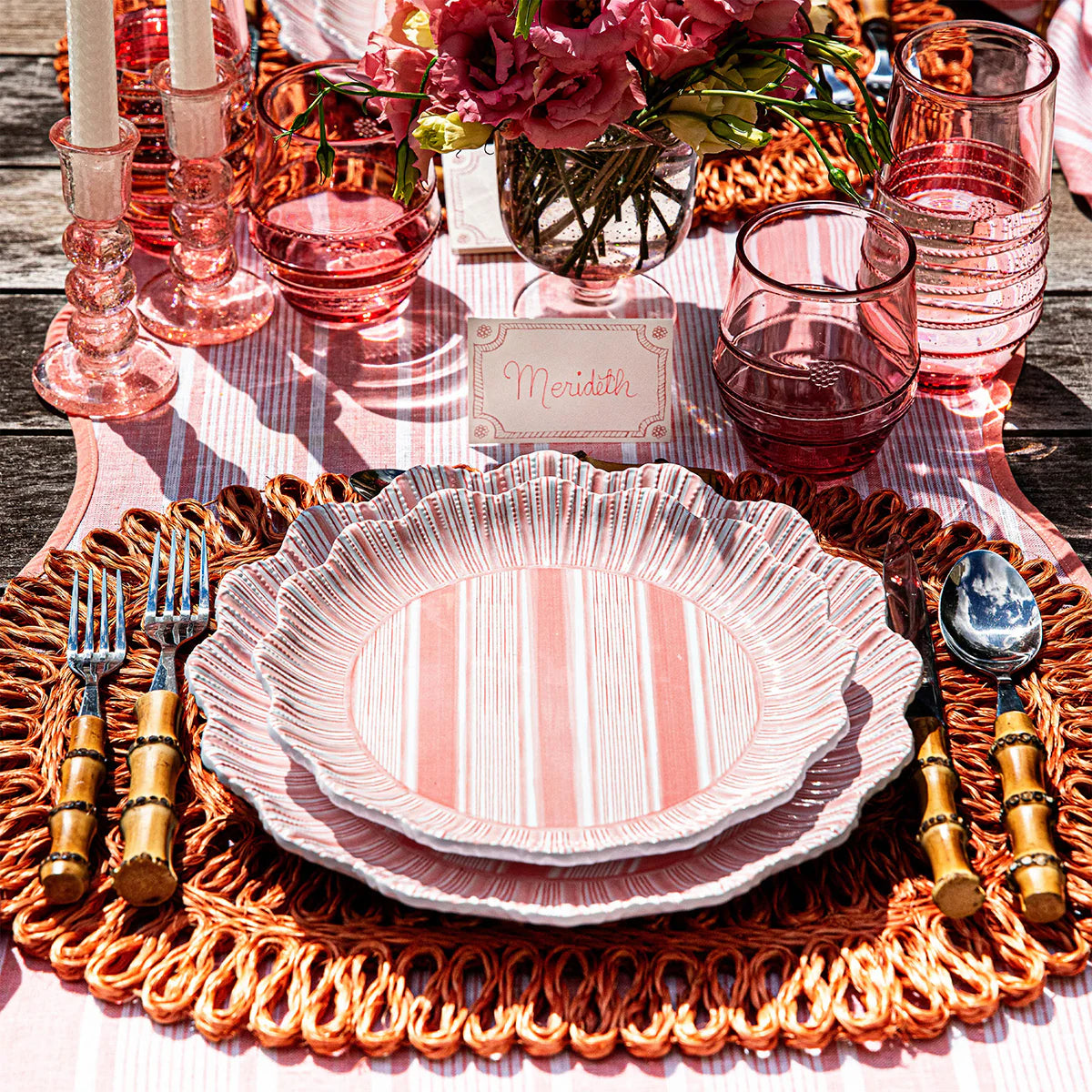 A vibrant outdoor table features Juliska’s Cabana Stripe Melamine Dinnerware Set of 4 in Coral, gold bamboo flatware, pink glassware, a woven charger, pink candles, flowers in a vase, and a name card labeled Meridith above the plates.