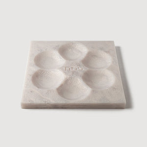 The Resin Seder Plate - NEW! by CDMX / BRUCI is a square, light-toned plate with six round indentations in a hexagonal pattern. Handcrafted in Mexico, its textured design includes a center embossed nub—perfect for Passover 2025.