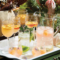 A tray holds the Juliska Isabella Acrylic Pitcher with a pink drink, lemon and peach slices, plus glasses garnished with herbs, citrus, and edible flowers—perfect for outdoor entertaining amid tropical decor.