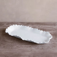 The Beatriz Ball Vida Colette Melamine Platter White - Large features elegant scalloped edges, resting on a brown textured surface against a beige wall—ideal for stylish presentations at any gathering.