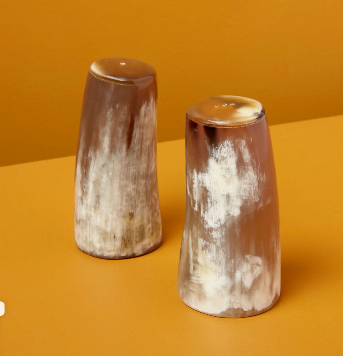 The Be Home Tusk Salt & Pepper Shaker - Large features a sleek brown and white marbled design inspired by tusks, displayed on an orange surface with an orange background—distinctive serveware to elevate your table.