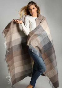 A woman in a white long-sleeve top and blue jeans wraps herself in the Alicia Peru Home Sloane Alpaca Throw, featuring superfine alpaca with tassel trim in brown, beige, and gray, against a plain gray background.
