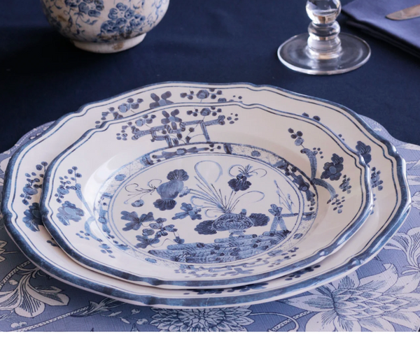 The Beatriz Ball Faenza Melamine Dinnerware Blue & White - Set of 4 features stacked plates with intricate blue and white patterns, styled with a matching placemat, clear wine glass, and blue-patterned bowl.