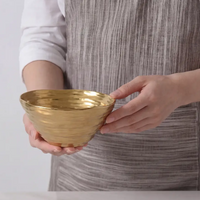 A person in a gray striped apron holds the Dorrato Gold Bowl - by PAMPA BAY, which is matte gold and dishwasher safe, standing behind a white surface.
