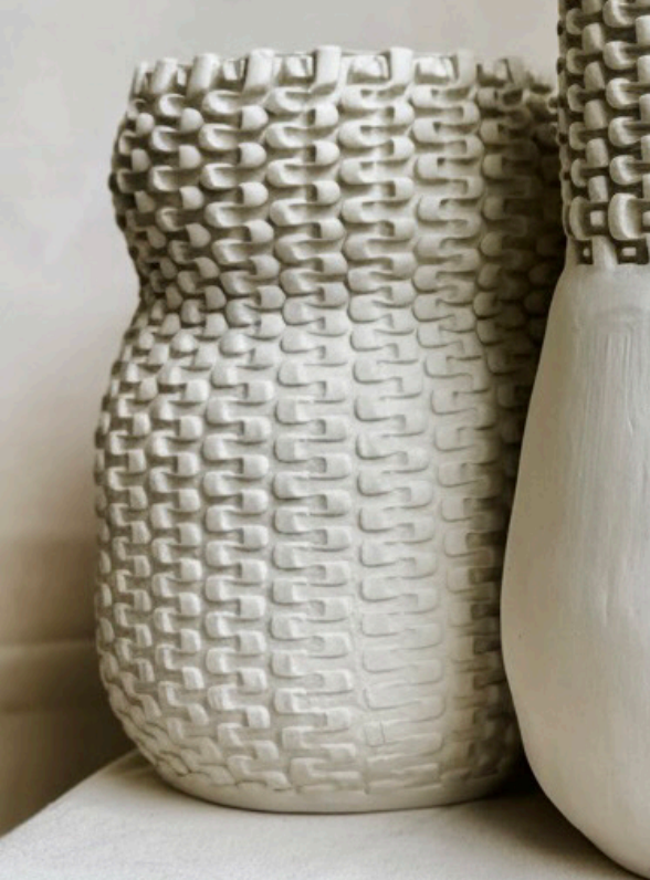 A cream-colored Chain Curve Vase by CYM Warkov Ceramics, featuring a textured, woven pattern and a wavy rim, sits beside another smooth unglazed porcelain vase from the Round Curvy Vase Collection, both serving as tasteful decorative accents on a light surface.