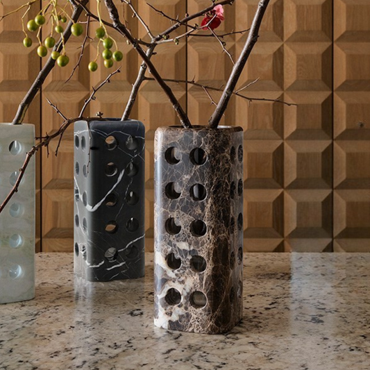 Three Geo Marble Vases by Greg Natale, each featuring a circular opening, stand on granite. The marble vases hold bare branches—some with green berries and one with a red flower—against a geometric wood wall backdrop.