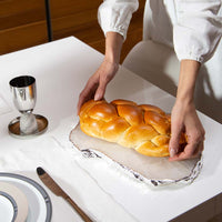 A person places a braided challah bread on the Kiva Large Platter - Crystal/Silver by Anna New York, set on a foil-covered table next to a pure silver-plated cup and an elegantly crafted challah board by the dining plate.