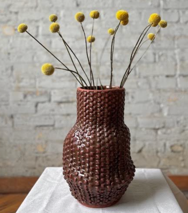 A textured, reddish-brown Serpent Vase - Red Iron by CYM Warkov Ceramics with a repeating grid pattern, holding a bouquet of yellow billy ball flowers for a touch of contemporary elegance.