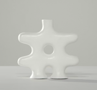 The Nedjma Vase by Schwung Design features a handcrafted ceramic form with a central hole and four rounded protrusions, displayed on a light gray surface against a plain gray background for an artisan-made accent.