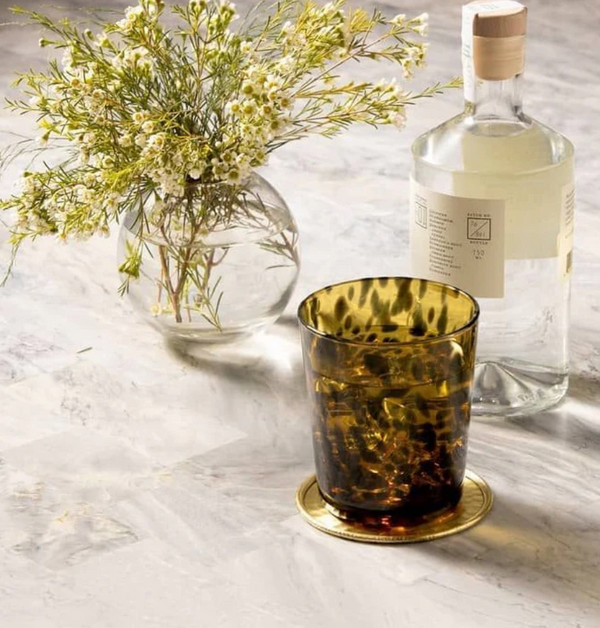 A round vase with small white flowers, a clear glass bottle with a wooden cap, and a Blue Pheasant Andrew Drinkware DOF - Tortoise glass filled with ice rest on a light marble surface.