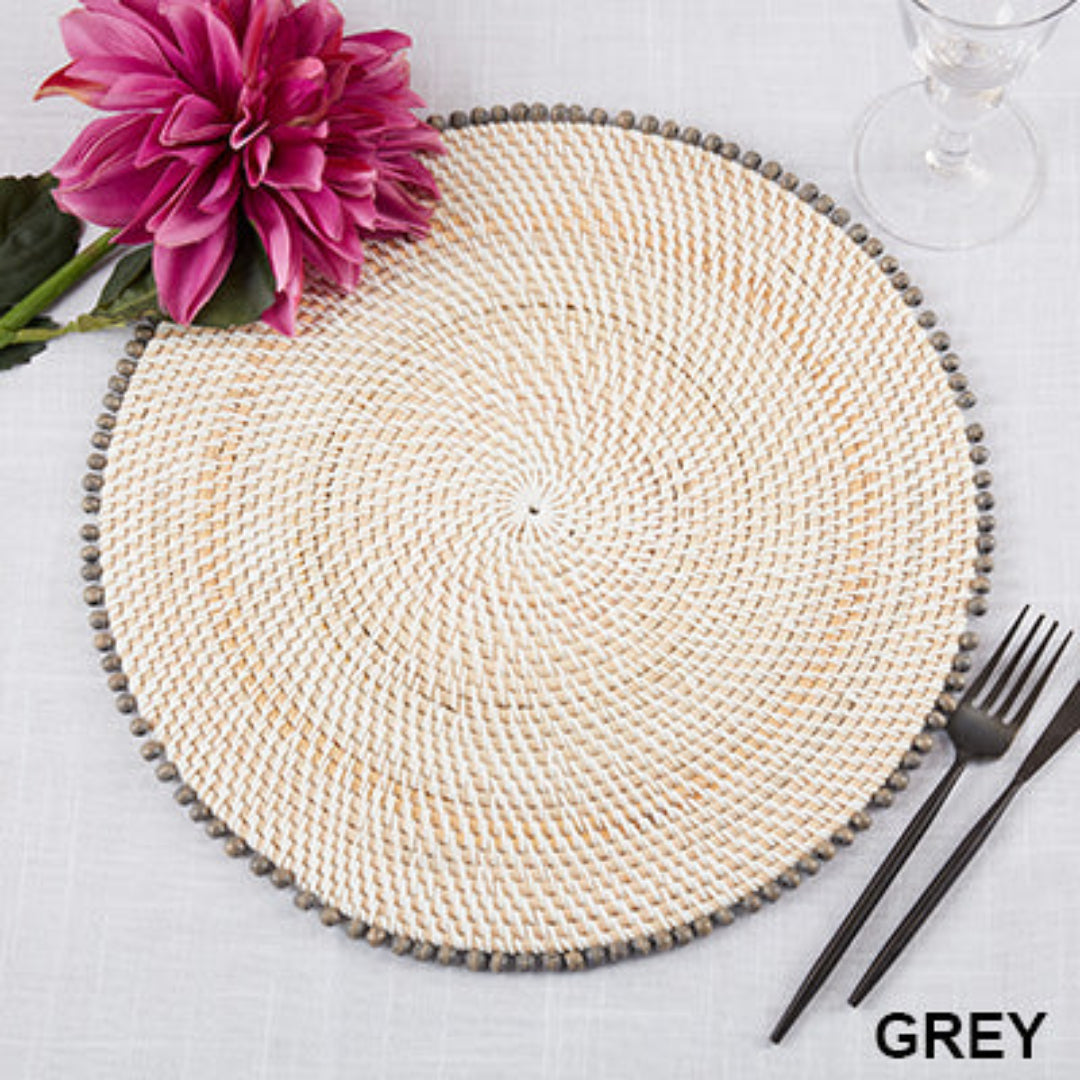 A Beaded Edge Round Placemat Set of 4 in Grey by Saro Trading rests on a white tablecloth, styled with a pink flower, glass, and black cutlery. “GREY” is noted in the bottom right corner for color reference.