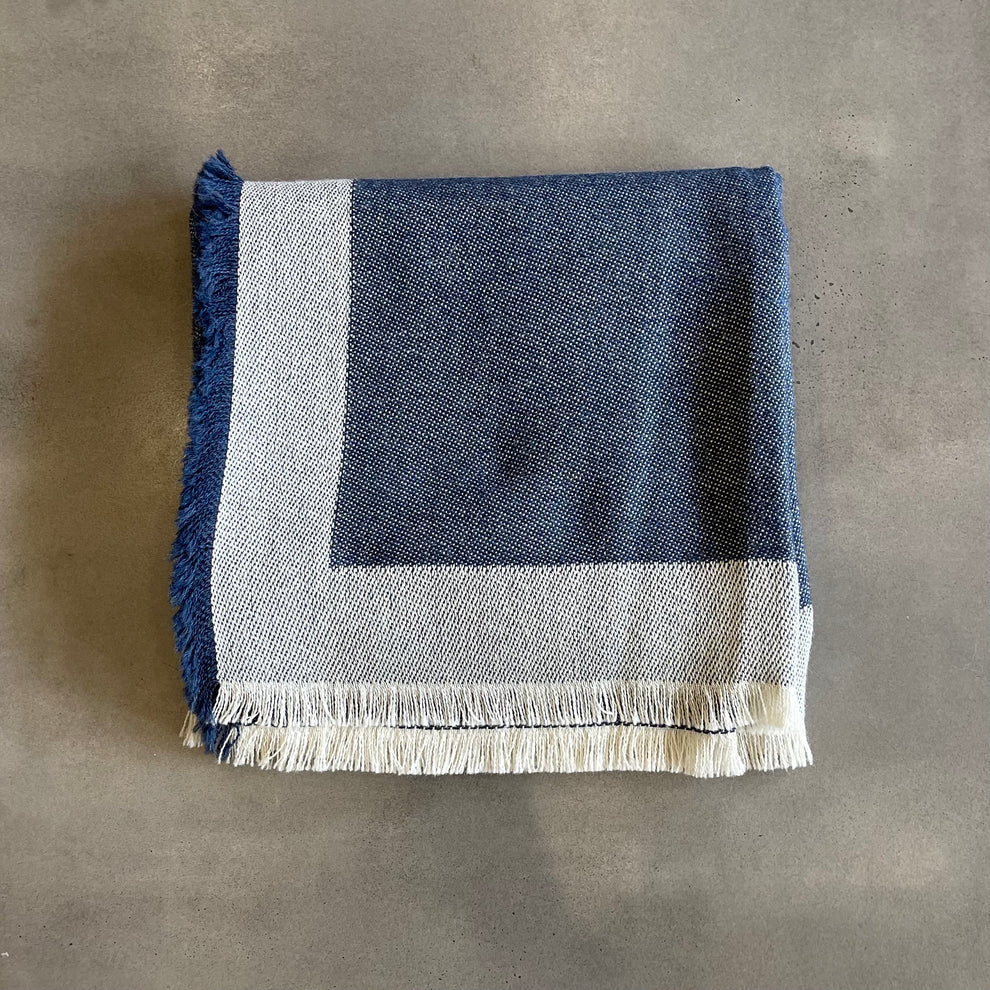 Alicia Peru Home’s Eyelash Alpaca Throw, featuring blue and white hues with delicate fringe, is folded neatly on a smooth gray surface.