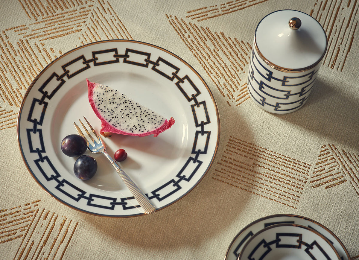 A plate from the Ginori 1735 Catene Nero Dinnerware collection, holding a slice of dragon fruit, two grapes, and a red berry, sits on a patterned tablecloth beside a matching lidded ceramic jar. A fork rests on the plate.