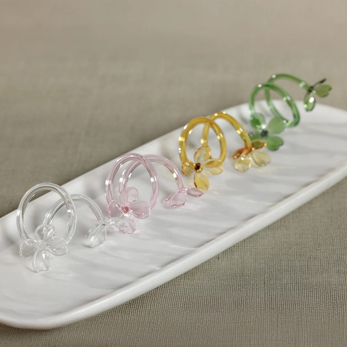 Four Botanica Napkin Rings by Zodax, each crafted from glass in distinct shades of white, pink, yellow, and green and adorned with delicate floral designs, are showcased upright on a white rectangular ceramic tray atop a beige fabric surface. These sculptural napkin rings bring an artistic flair to your decorative tableware collection.