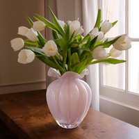 The Beatriz Ball Glass Vento Vase Camellia - Large, in a pale pink hue and filled with white tulips and green leaves, sits on a wooden table near a window with white curtains that let in natural light.