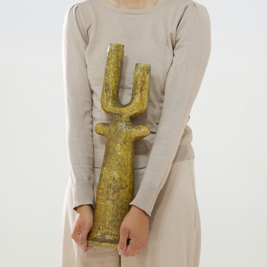 A person in a beige outfit holds the Govad Candleholder 20″ by Schwung Design—a large, gold, U-shaped sculpture with both hands. The candleholder’s two raised arms evoke the elegance of a classic candlestick and reflect the intricate essence of Moroccan artistry.