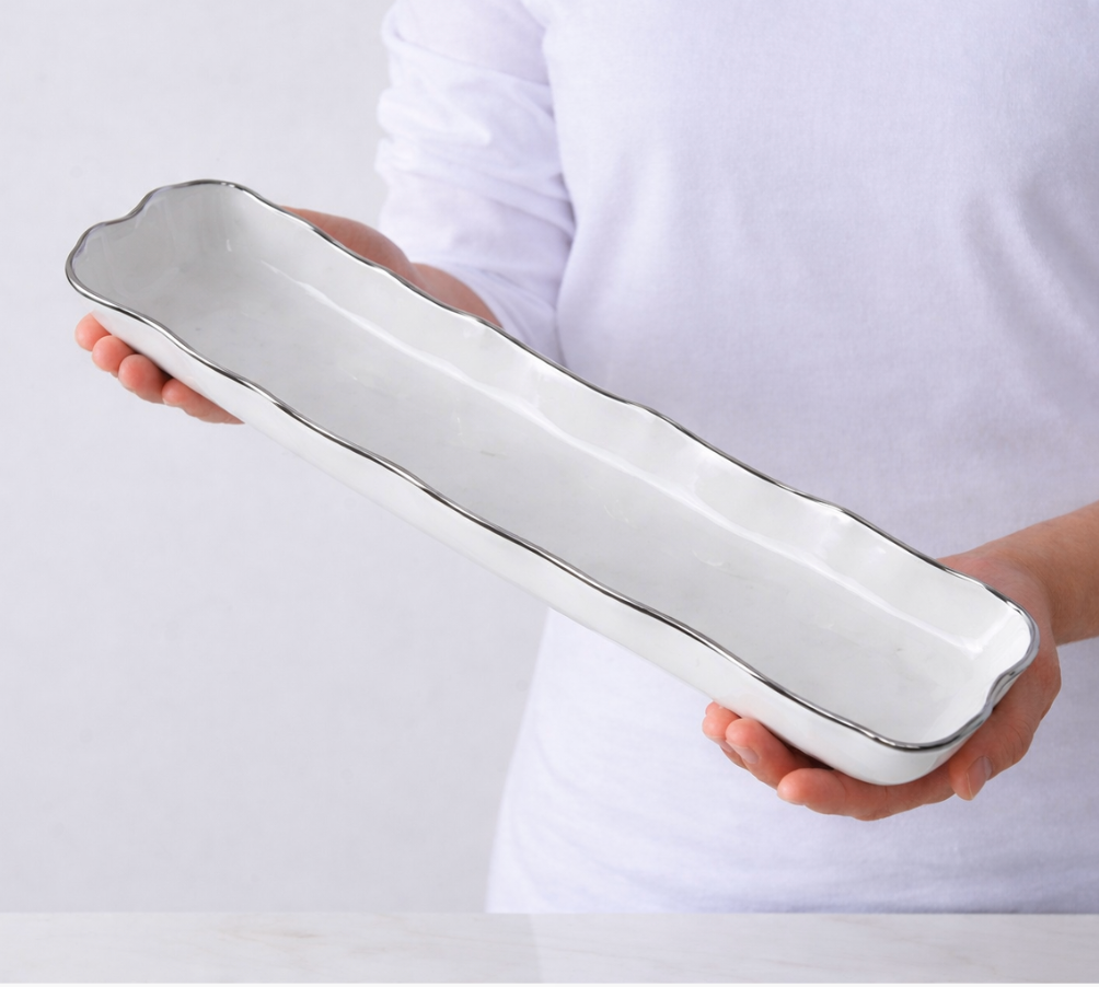 A person in a white shirt holds the PAMPA BAY Portofino Plata Cracker Tray, a long, rectangular white ceramic serving dish with wavy edges and a subtle metallic rim.