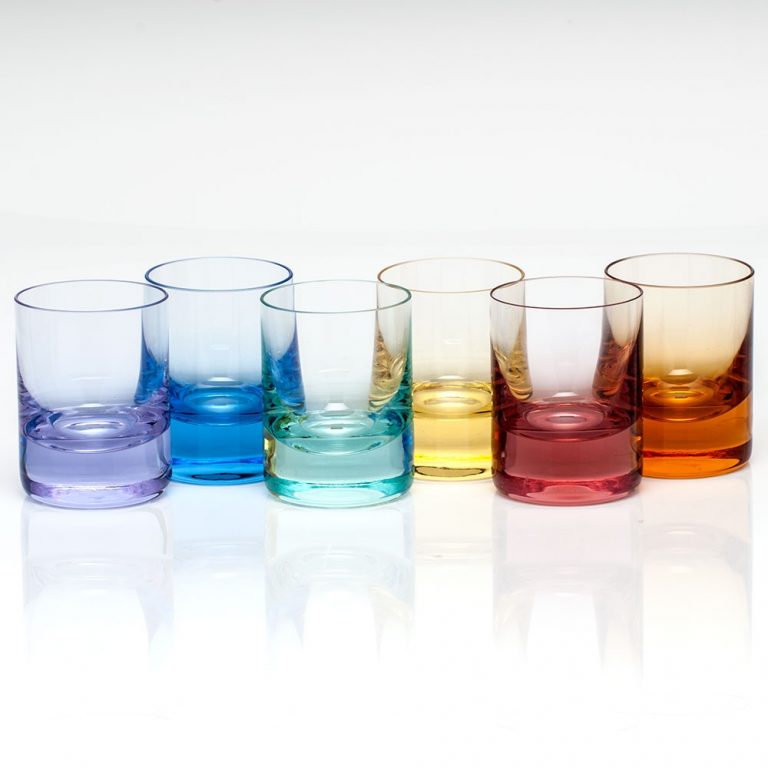 A row of six sleek, hand-blown Whiskey Set Shot Glasses by Moser, each in a distinct transparent color—purple, blue, light blue, yellow, pink, and orange—showcasing vibrant Moser artistry. The glasses are arranged against a white background with a reflective surface that highlights their elegant lines and colorful brilliance.