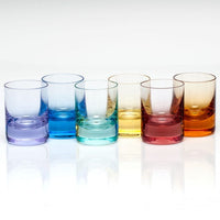 A row of six sleek, hand-blown Whiskey Set Shot Glasses by Moser, each in a distinct transparent color—purple, blue, light blue, yellow, pink, and orange—showcasing vibrant Moser artistry. The glasses are arranged against a white background with a reflective surface that highlights their elegant lines and colorful brilliance.