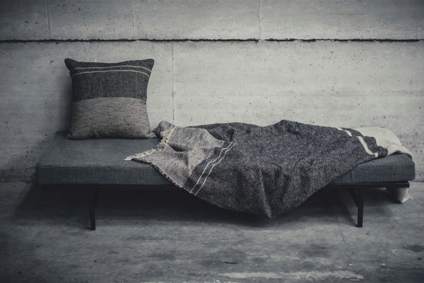 A minimalist gray daybed with a Libeco Home Francis Pillow 25 x 25 and a textured gray blanket sits against a bare concrete wall in an industrial setting.