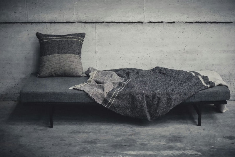 A minimalist gray daybed with a Libeco Home Francis Pillow 25 x 25 and a textured gray blanket sits against a bare concrete wall in an industrial setting.