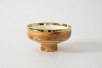 The Madera Footed Bowl by PAMPA BAY is a round bowl with a wood grain pattern and shiny gold accents, featuring a wide rim and a short, cylindrical pedestal base, set against a plain white background.