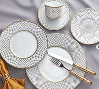 The Mottahedeh/Robert Haviland Home Dinnerware-Gold set features patterned plates, a cup and saucer, gold-handled flatware, and a beaded napkin ring atop a light gray tablecloth for an elegant dining setting.