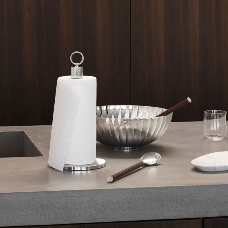 A paper towel roll on a metal holder, a silver bowl with chopsticks, a glass, the Georg Jensen Bernadotte Stainless and Wood Salad Set spoon, and a small dish are arranged on a modern gray kitchen countertop. Dark wooden cabinets are in the background.