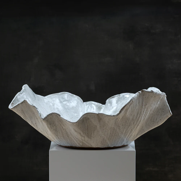 The Happenstance II Centerpiece Bowl by Kirti Sheoran Studio, a large white ceramic piece crafted from stoneware clay with wavy, uneven edges, sits atop a white pedestal against a dark, textured background. This one-of-a-kind bowl is perfect for anyone seeking a statement centerpiece.