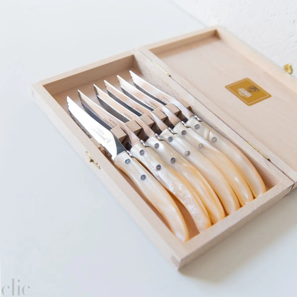 A wooden box holds six Claude Dozorme Berlingot Steak Knives in assorted colors, each with a gleaming stainless steel blade and a curved, pearlescent handle, all neatly arranged in individual slots inside the open case.