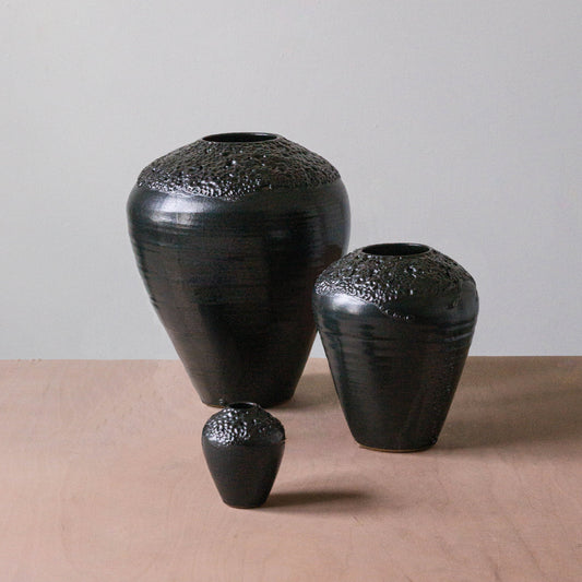 The Esme Crawl Vase - Black Lava by Kalya OD Studio features large and medium water-safe vases with black textured finishes, perfect for fresh or dried arrangements on any light brown surface against a gray wall.
