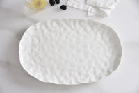 The PAMPA BAY Willow White Platter Oval Large, featuring a textured ceramic finish, rests on a white surface beside a folded napkin, a glass with lemon, and several blackberries.