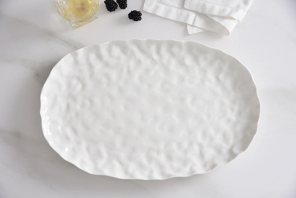 The PAMPA BAY Willow White Platter Oval Large, featuring a textured ceramic finish, rests on a white surface beside a folded napkin, a glass with lemon, and several blackberries.