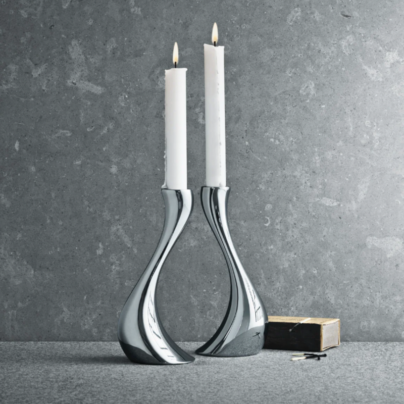 Two tall white candles glow in the Georg Jensen Cobra Candleholder 2 Piece Set, displayed on a gray stone surface with a matchbox and matches—ideal for adding a modern touch to minimalist home decor.