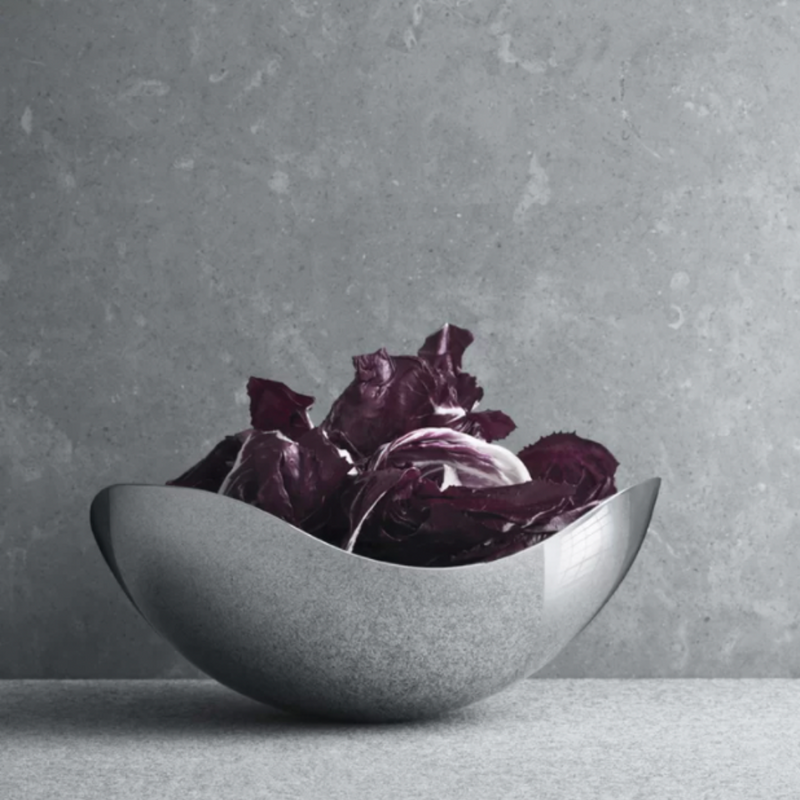 The Bloom Serving Bowl by Georg Jensen, with its modern curved stainless steel design and mirror-polished finish, elegantly cradles deep purple radicchio on a gray surface set against a textured gray backdrop.