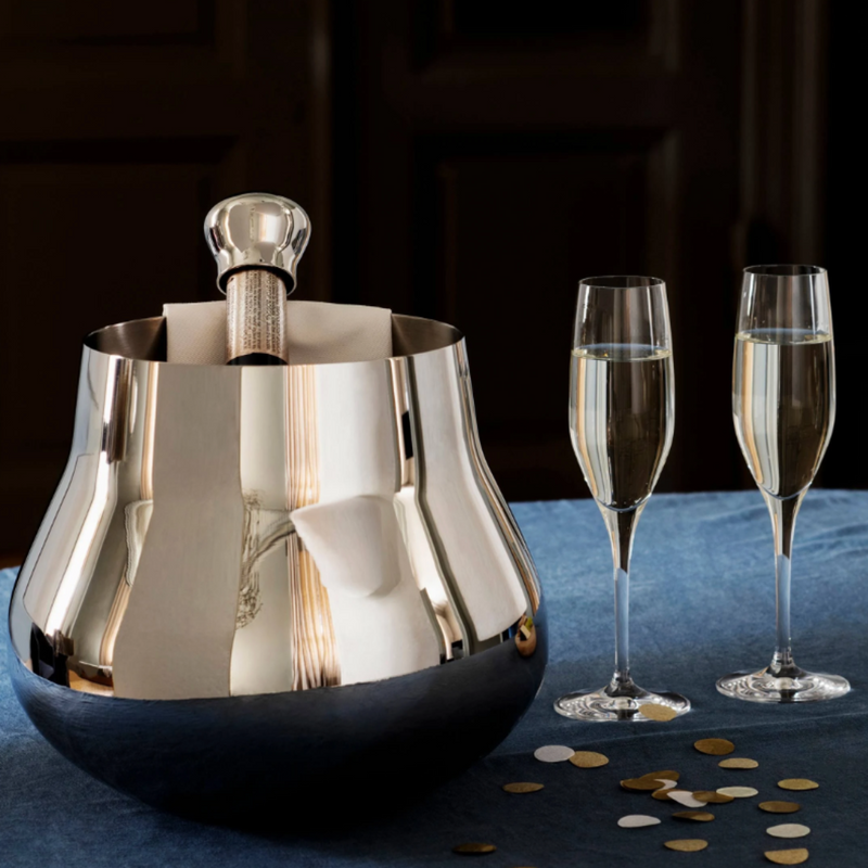 The Georg Jensen Sky Champagne & Wine Cooler, holding a bottle of champagne and napkin, rests on a blue tablecloth beside two filled flutes and scattered gold and white confetti.