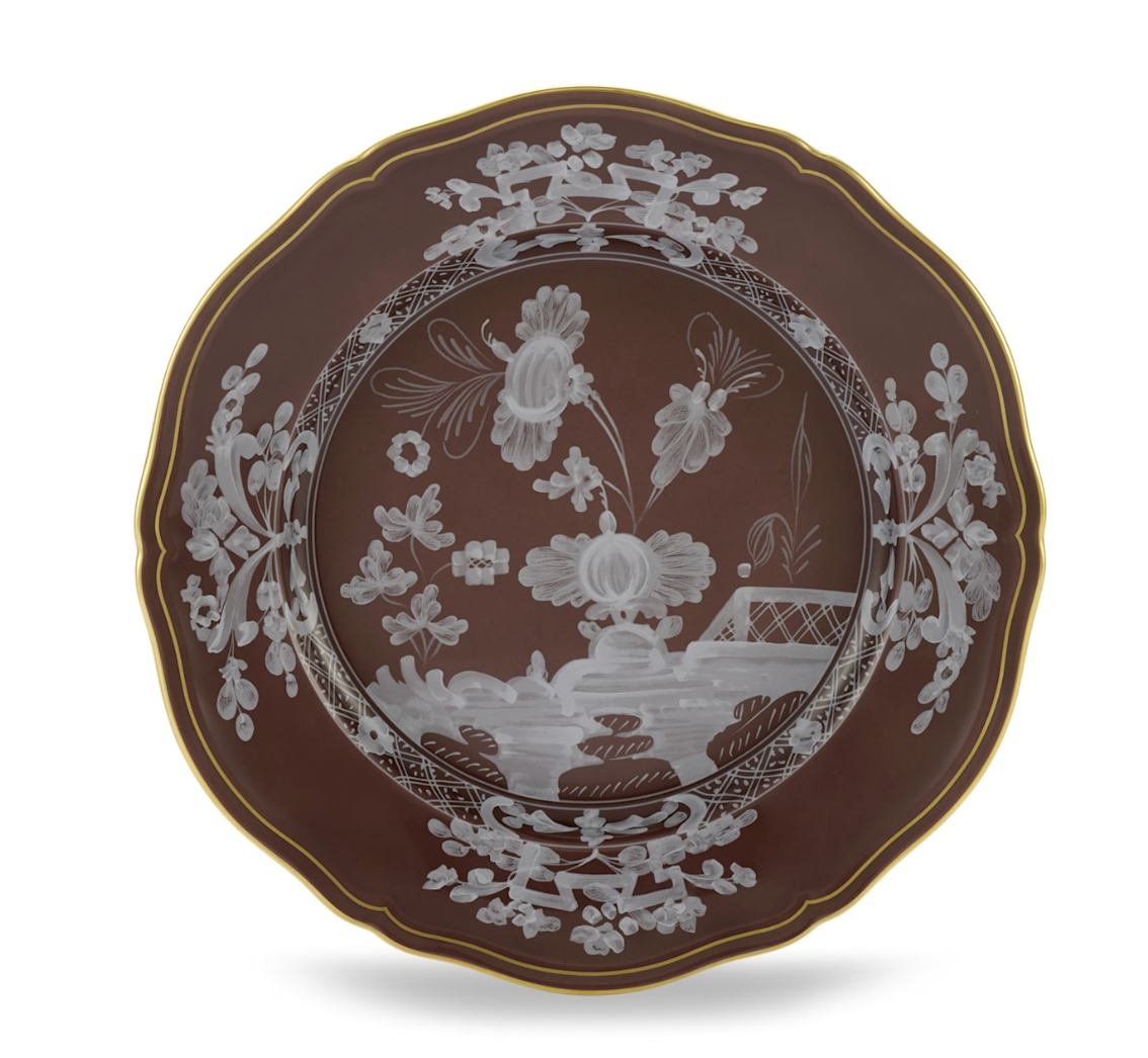 The Oriente Italiano Castagna plate by Ginori 1735 is a round, brown porcelain piece with scalloped edges and a gold rim, adorned with intricate white floral and landscape motifs. Made in Italy.