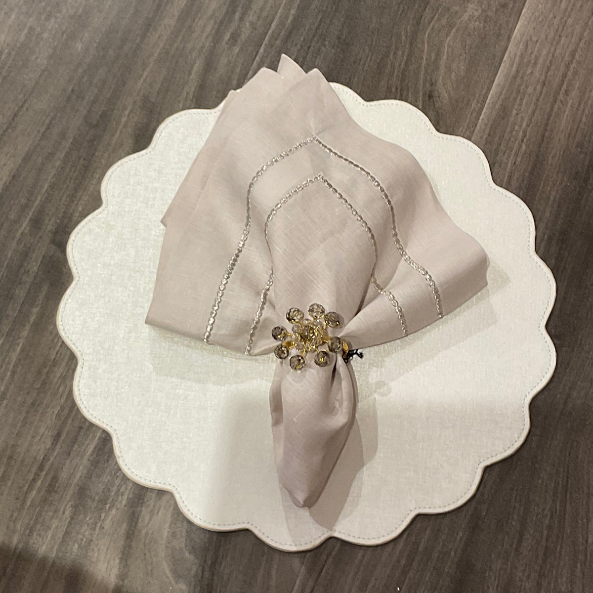 A pale folded napkin with a gold beaded ring sits on a Deborah Rhodes Victoria Round Scallop Placemat Set of 4, adding easy-care elegance to the wooden table.