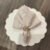 A pale folded napkin with a gold beaded ring sits on a Deborah Rhodes Victoria Round Scallop Placemat Set of 4, adding easy-care elegance to the wooden table.