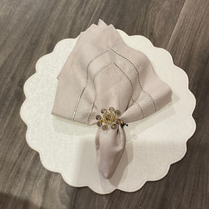 A pale folded napkin with a gold beaded ring sits on a Deborah Rhodes Victoria Round Scallop Placemat Set of 4, adding easy-care elegance to the wooden table.
