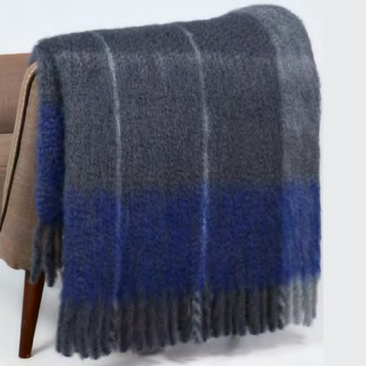 The Mantas Ezcaray Twist Frame Throw, a fuzzy mohair blanket handcrafted in Spain with blue and gray plaid and fringed edges, is draped over a brown chair.