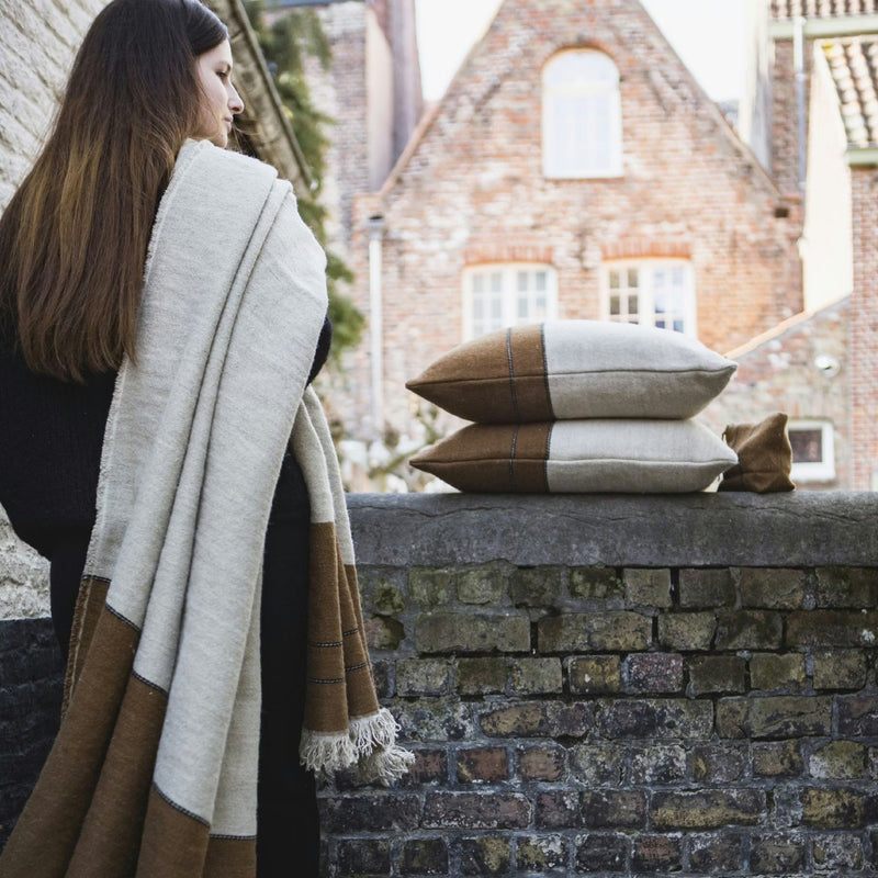 A woman with long brown hair drapes a Libeco Home Gus Throw over her shoulder near a brick wall. Nearby, three linen wool pillows with natural stripes are stacked with old brick buildings visible in the background.