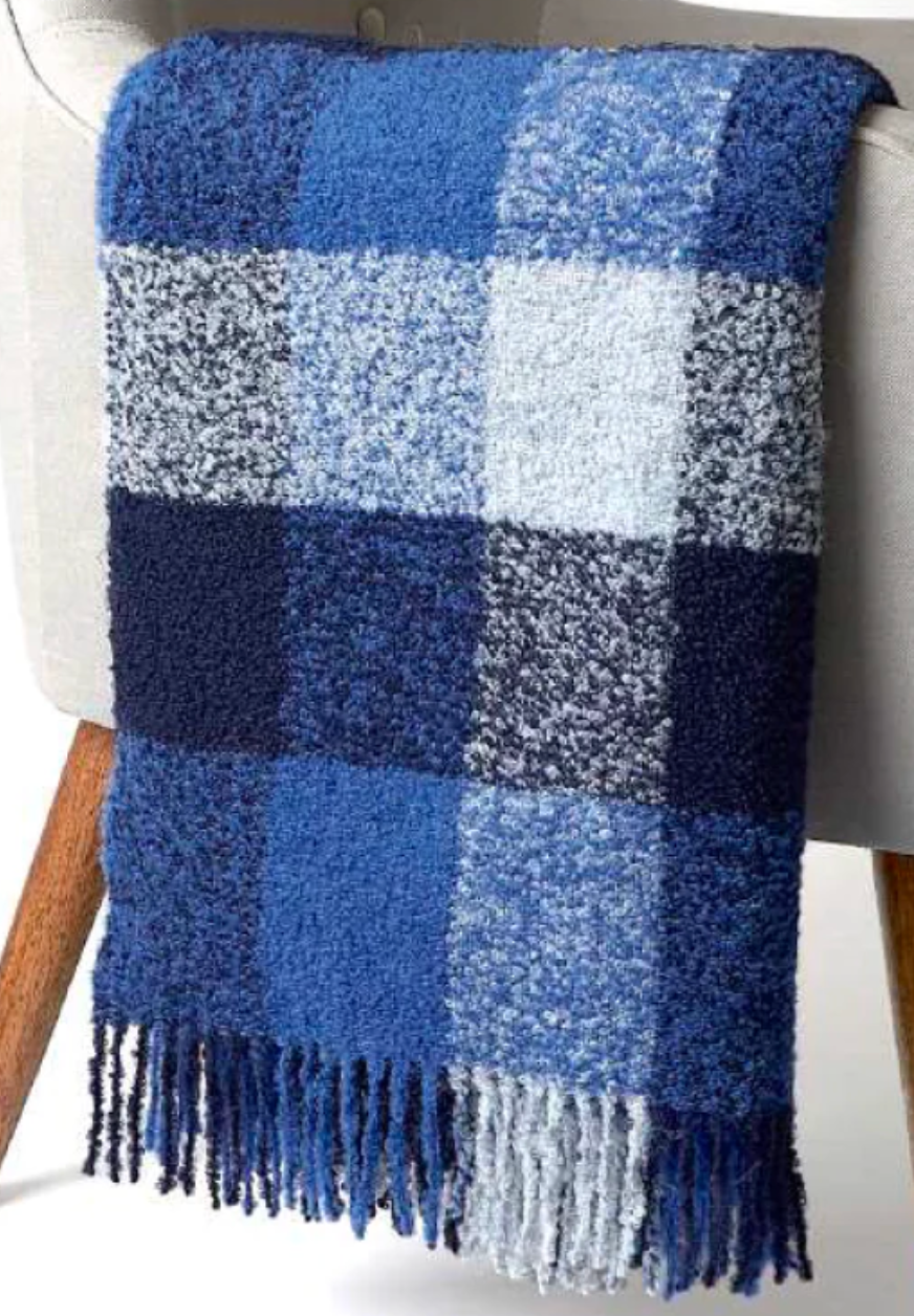 Alicia Peru Home’s Bristol Boucle Throw in Cobalt / Blue, featuring textured squares in various shades of blue and finished with fringed ends, is draped over the back of a light-colored chair.