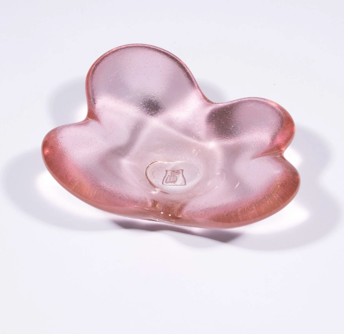The Cloud Dish by Tyler Barry Design is a hand-blown, pink translucent glass bowl with three rounded lobes and an organic shape. Displayed on a white background, it features a small emblem at the center for added detail.