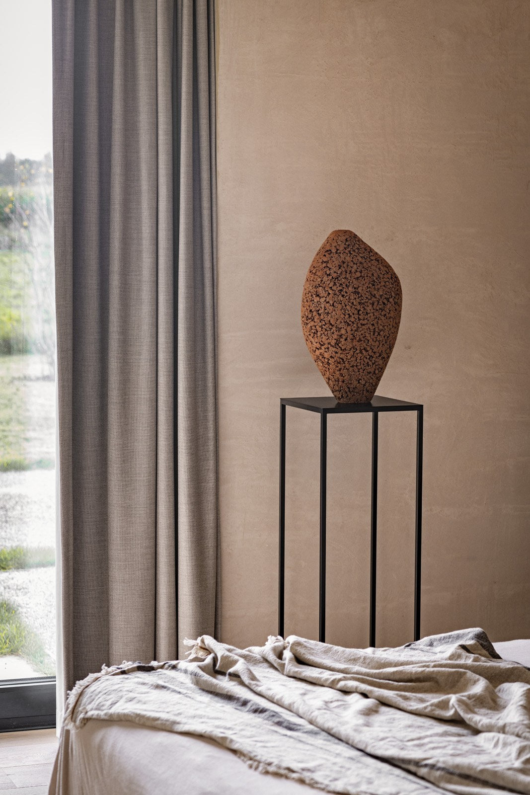 A tall, black metal stand displays the Gardeco Formigueiro Vase 5 in brown natural next to beige curtains and a window. In the foreground, a light-colored bed with a crumpled blanket completes the setting.
