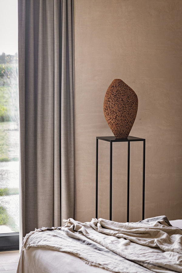 A tall, black metal stand displays the Gardeco Formigueiro Vase 5 in brown natural next to beige curtains and a window. In the foreground, a light-colored bed with a crumpled blanket completes the setting.