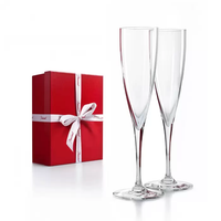 Two empty Dom Perignon Champagne Flutes by Baccarat stand side by side next to a closed red gift box tied with a white ribbon, all set against a plain white background.