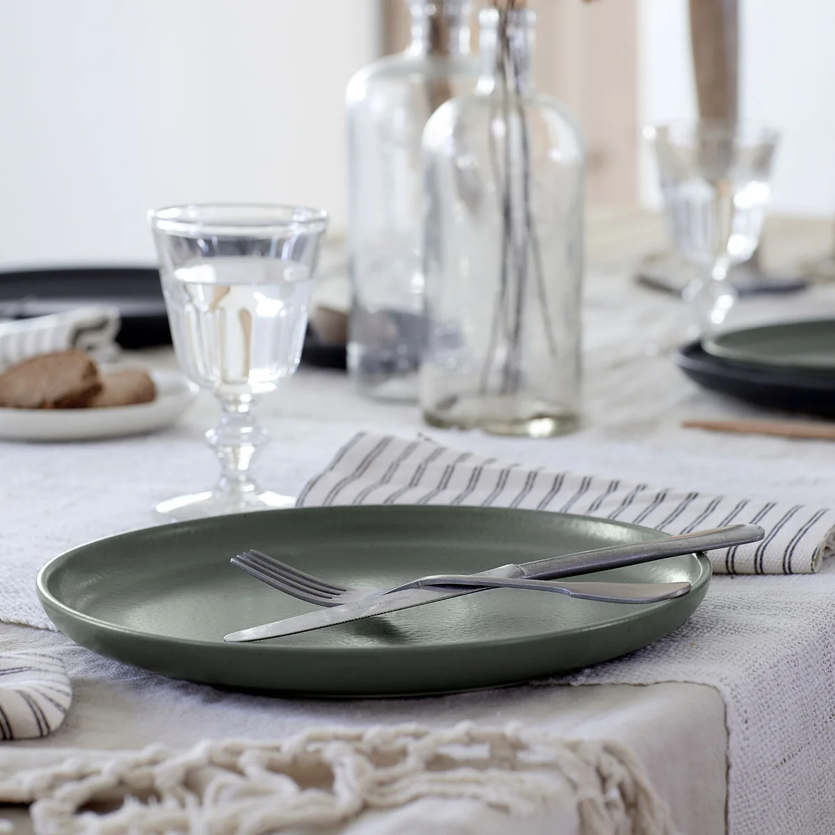 A Pacifica Dinnerware Artichoke plate from Casafina/Costa Nova, accompanied by a fork and knife, rests elegantly on a set table with a glass of water, striped napkin, and glass bottles atop a textured white tablecloth—a beautiful showcase of luxury tableware.