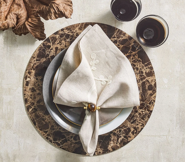 Table setting with a marble-patterned charger, gray and white plates, a beige embroidered napkin accented by the Kim Seybert Mineral Napkin Ring set of 4 in Brown, and two glasses of a dark beverage, viewed from above on a light surface.