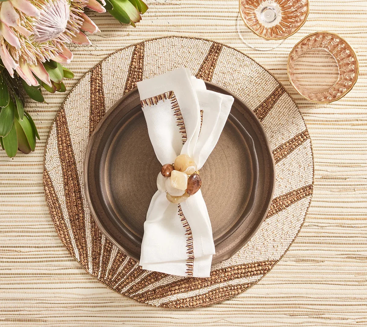 A brown plate is topped with a Kim Seybert Filament Napkin in white and brown, elegantly held by a beaded napkin ring and set on a woven placemat with brown rays. Pink flowers and two glasses are positioned at the top left and right, completing this sophisticated tablescape featuring the Filament Napkin Set of 4 by Kim Seybert.